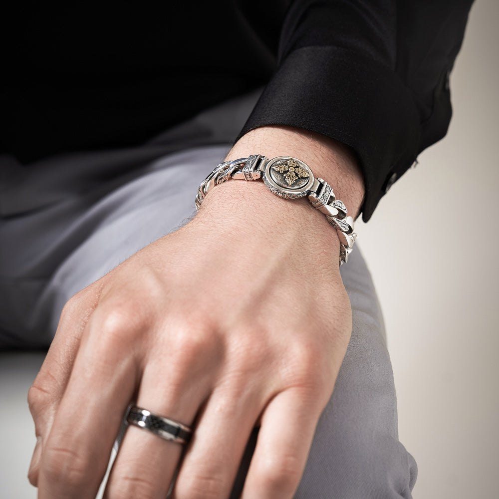 Close-up of a man’s wrist resting on his knee, wearing the Ronatelli Zeus 11 mm curb 925 sterling silver bracelet with a gold-accented double vajra medallion